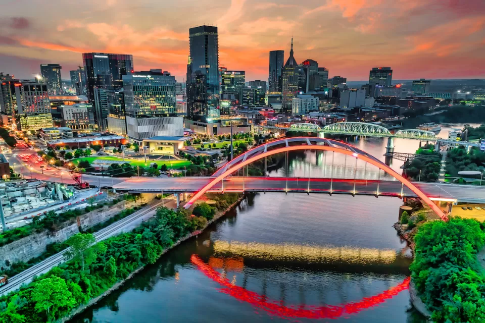 Aerial view of Nashville, Tennessee skyline with river and bridge in foreground against sunrise or sunset sky.